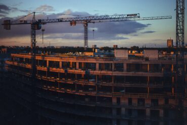 Строящаяся многоэтажная бетонная конструкция с двумя башенными кранами на фоне вечернего неба